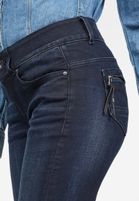 Jeans de mezclilla azul oscuro con una cintura alta, corte slim, bolsillos delanteros y un bolsillo con cremallera en la cadera derecha. Textura suave.