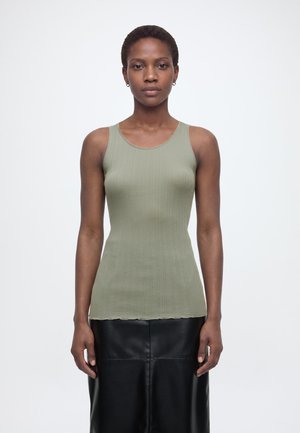 Staande persoon draagt een mouwloos geribd lichtgroen topje en een zwarte leren rok, kijkend naar voren tegen een effen witte achtergrond.