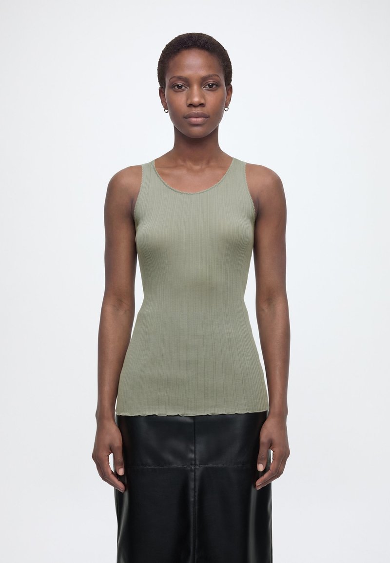 Άτομο που στέκεται φορώντας ένα αμάνικο ριμπ πλεκτό ανοιχτοπράσινο τοπ και μαύρη δερμάτινη φούστα, κοιτάζοντας μπροστά με φόντο ένα απλό λευκό φόντο.