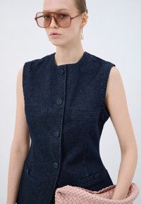 Chaleco de mezclilla sin mangas en azul oscuro con un cuello redondo, botones delanteros y dos bolsillos. Sosteniendo un bolso de mano de mimbre rosa.