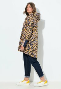 Imperméable à capuche avec un fond marine, un motif floral jaune et des manches longues. Associé à un jeans en denim foncé et des chaussures blanches avec des accents jaunes.