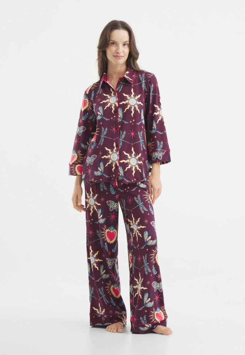 Paarse pyjama set met een knoopsluiting bovenkant en wijde broekspijpen, versierd met zonnen, harten en bloemenpatronen in felle kleuren. Zachte stoffen textuur.