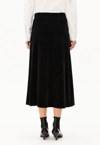 Schwarzer Samt-Midi-Rock mit glatter Textur, ausgestellt geschnitten und mit einem Taillenbund. Kombiniert mit spitzen schwarzen Stiefeletten.