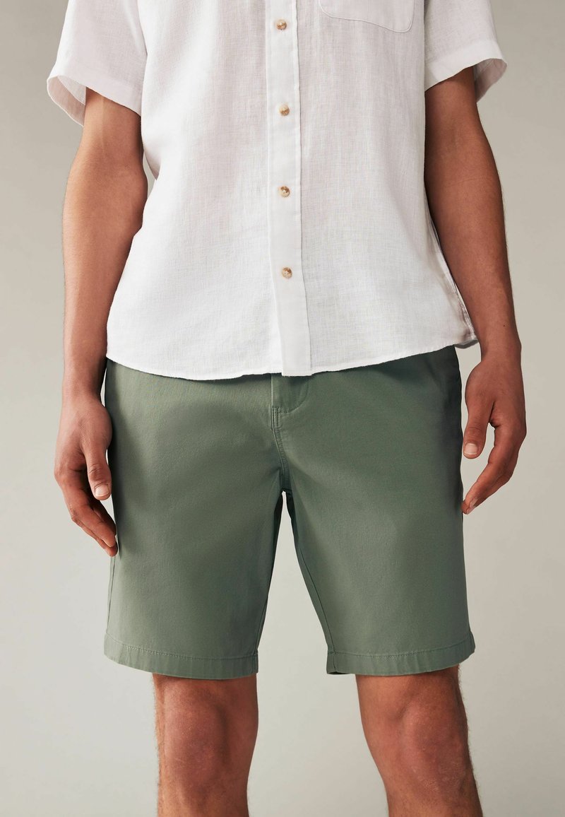 Leichtes weißes Leinenhemd mit kurzen Ärmeln und einer Brusttasche. Kombiniert mit dunkelgrünen Shorts, die ein einfaches Design und umgeschlagene Bündchen haben.