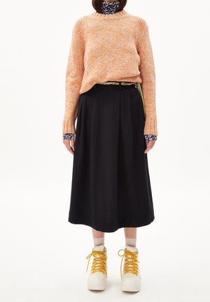Person wearing an orange knit sweater over floral turtleneck, black midi skirt, beige socks, and white boots with yellow laces standing against white.