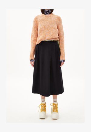 Personne portant un pull en tricot orange sur un col roulé à motif floral, une jupe midi noire, des chaussettes beiges et des bottes blanches avec des lacets jaunes, debout devant un fond blanc.