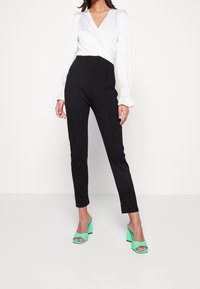 Vit satinblus med lång puffärm, parat med svarta figurnära byxor; modellen bär gröna klackskor med öppen tå.