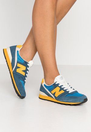 Benen met blauwe, gele en witte New Balance sneakers met witte veters, staand op een wit oppervlak tegen een effen achtergrond.