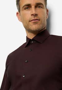 Burgunderfarbenes Hemd mit strukturiertem Muster, umlegekragen und schwarzen Knöpfen. Der Stoff wirkt glatt mit leichtem Glanz. Lässiges Design.