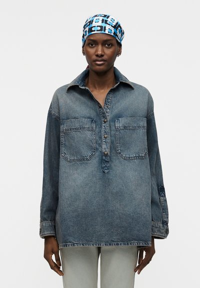 Femme portant une chemise en denim délavée et ample avec des poches avant et des boutons, un pantalon clair, et un foulard bleu à motifs, debout devant un fond uni.