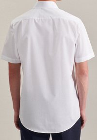 Bílá košile s krátkým rukávem a špičatým límcem, s zakulaceným lemem a hladkou texturou. Pohled zezadu ukazuje čisté linie a detaily švů.