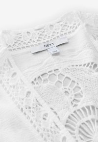Indumento bianco con dettagli in pizzo intricati e motivi geometrici. L'etichetta reca la scritta "NEXT". Tessuto morbido con una finitura delicata.
