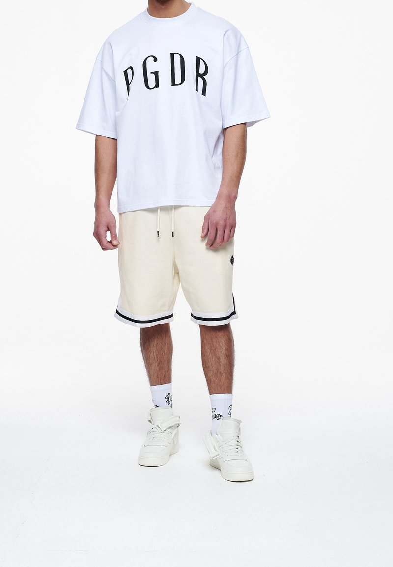 Biała bawełniana koszulka z czarnym napisem "PGDR", zestawiona z kremowymi szortami z czarnym wykończeniem oraz białymi trampkami w połączeniu ze skarpetkami w wzory.