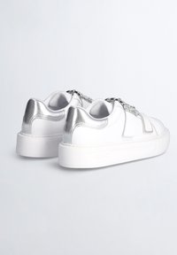 Hvide sneakers med sølvdetaljer, der har en glat læderoverdel, tyk hvid sål og dekorativ kædedetalje på forsiden.