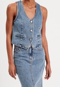 Veste en jean avec col en V, boutons argentés et deux poches avant. Teinture bleu clair et coupe courte. Portée avec une jupe en jean assortie.