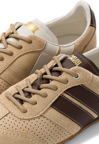 Une paire de baskets en daim beige avec des bandes en cuir marron, des lacets beiges et un logo doré sur la languette.