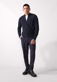 Jachetă tricotată cu fermoar navy, cu textură canelată, asociată cu pantaloni dark slim-fit și pantofi negri, stând în fața unui fundal neutru.