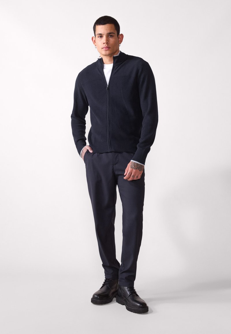 Veste tricot zippée bleu marine avec texture côtelée, associée à un pantalon slim foncé et des chaussures noires, se tenant devant un arrière-plan neutre.