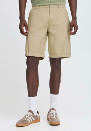 Beige khaki shorts met een opgerolde zoom, gemaakt van lichtgewicht katoen. Draagt witte sokken en bruin suède sneakers met crèmekleurige strepen.