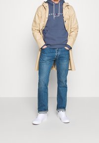 Impermeabile beige sopra un hoodie blu navy con logo bianco, jeans blu e sneakers bianche. L'outfit presenta un design rilassato e a strati.
