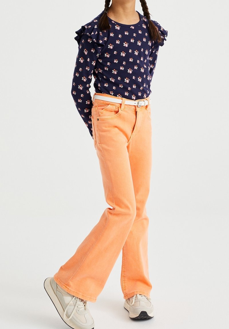 Jeune fille avec des cheveux tressés portant une chemise à manches longues bleu marine à motifs floraux, un pantalon évasé orange avec une ceinture blanche, et des baskets blanches.