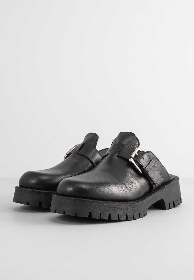 Sabots en cuir noir avec une épaisse semelle en caoutchouc, présentant une boucle et un design à dos ouvert. Texture lisse avec de subtils détails de couture.