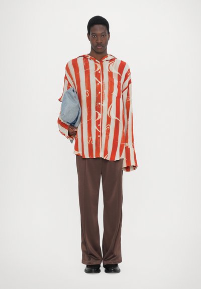 Red and white striped oversized shirt with a hood, paired with brown trousers. The individual holds a gray fabric bag. Smooth texture.