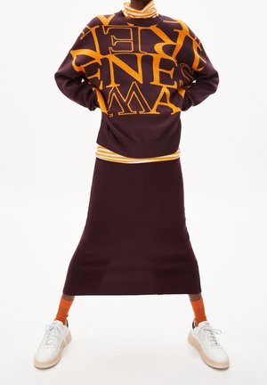 Lila Pullover mit großen gelben Buchstaben, kombiniert mit einem figurbetonten lila Rock. Weiße Sneakers und orangefarbene Socken runden das Outfit ab.