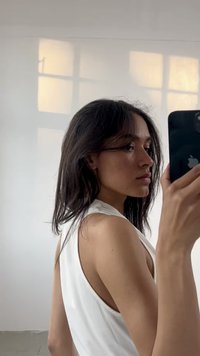 Portret osobe s tamnom, valovitom kosom, koja nosi bijelu majicu bez rukava, držeći crni pametni telefon na dobro osvijetljenoj, neutralnoj pozadini.