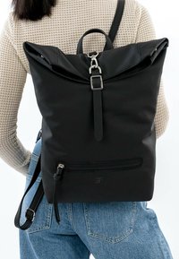 Schwarzer Rolltop-Rucksack aus strukturiertem Stoff, mit silbernen Beschlägen, verstellbaren Trägern und einer vorderen Reißverschlusstasche zur Aufbewahrung.