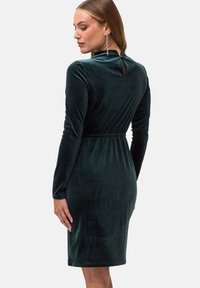 Ciemnozielona welurowa sukienka z długimi rękawami, dopasowanym wcięciem w talii i detalem w formie otworu na plecach. Gładka faktura i długość do kolan.