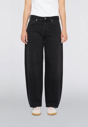Persona che indossa jeans di denim neri larghi, un top bianco e sneakers bianche con strisce azzurre, in piedi davanti a uno sfondo semplice.