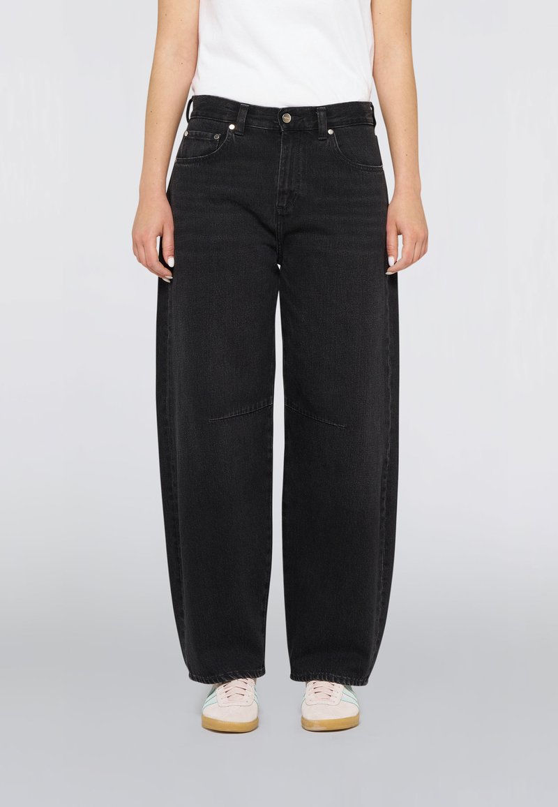 Personne portant un jean denim noir ample, un haut blanc et des baskets blanches avec des rayures bleu clair, debout devant un fond uni.