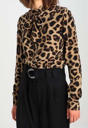 Frau trägt eine langärmelige Bluse mit Leopardenmuster und Schleifenkragen, die in eine hoch taillierte schwarze Hose mit einem Doppelring-Gürtel gesteckt ist.