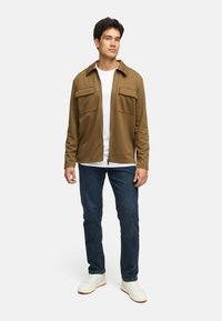 Brun zip-up skjortejakke med to brystlommer, iført over en hvid t-shirt. Parret med blå jeans og hvide sneakers.