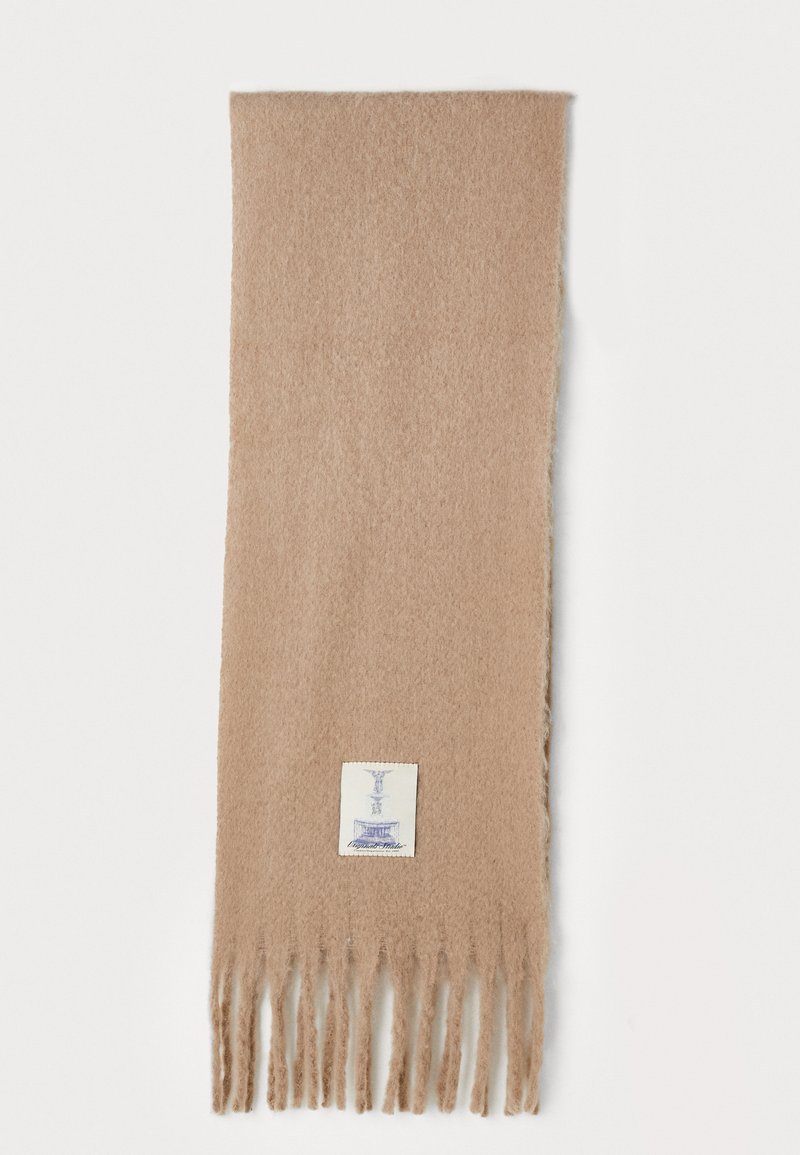 Lys beige skjerf med myk tekstur, med frynsekant og en liten etikett med blå tekst på hvit bakgrunn.