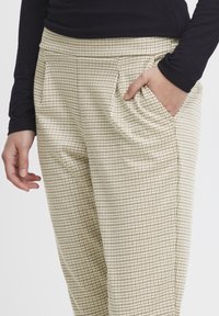 Beige karierte Hose aus strukturiertem Stoff, mit Seitentaschen und einer entspannten Passform. Kombiniert mit einem dunklen Langarmoberteil.