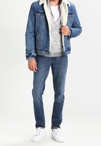 Veste en denim avec col en fausse fourrure, sweat à capuche gris zippé, T-shirt blanc, jeans bleus et baskets blanches. Tenue décontractée en couches.