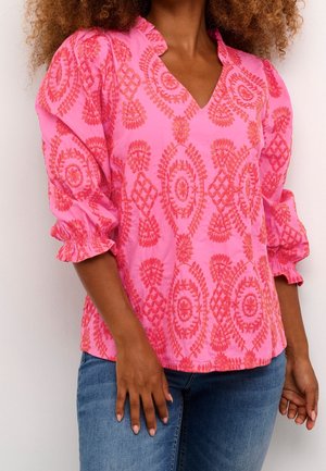 Frau, die eine pinke Bluse mit roter Blumenstickerei, gerafften Dreiviertelärmeln und blauen Jeans vor einem einfarbigen hellen Hintergrund trägt.