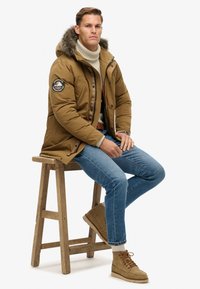 Chaqueta acolchada marrón con capucha forrada de piel sintética, cierre con cremallera y parche en el brazo izquierdo. Lleva puesta sobre un suéter beige, combinado con jeans azules y botas marrones.