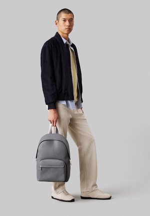 Homme portant un pantalon beige, une veste bleu marine et une chemise rayée, tenant un sac à dos en cuir gris par la poignée sur un fond gris clair.