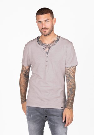Camisetta henley a maniche corte color beige chiaro con tre bottoni, morbida al tatto e dal taglio rilassato. Indossata con jeans grigi sbiaditi, che mostrano tatuaggi dettagliati.