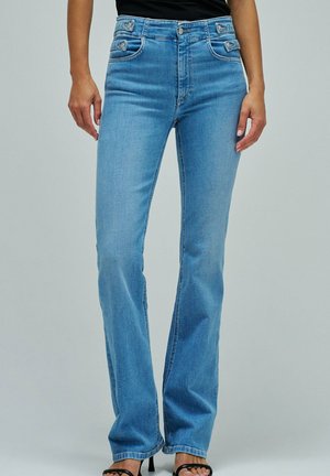 Femme portant un jean droit taille haute bleu clair et des talons ouverts noirs, debout devant un fond uni.