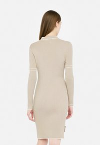 Abito beige a costine con maniche lunghe, orlo curvo e dettagli in bianco. Presenta una silhouette aderente e uno spacco laterale.