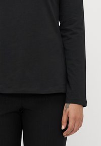 Person wearing a black long-sleeve shirt and black pants, showing a hand with a small key-shaped tattoo on the finger.