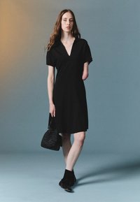 Robe noire à manches courtes avec un décolleté en V. Le modèle tient un petit sac à main noir texturé, portant des bottines noires et des chaussettes. Design simple.