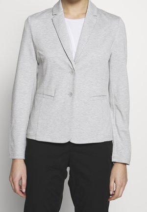 Frau trägt einen hellgrauen, taillierten Blazer mit zwei Knöpfen, Vordertaschen, über einem weißen Hemd und schwarzen Hosen, stehend vor einem schlichten Hintergrund.