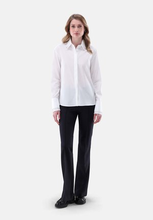 Camicia bianca con colletto classico, abbinata a pantaloni neri a palazzo e scarpe nere flat, su uno sfondo neutro.