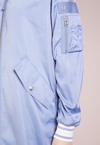 Chaqueta bomber azul claro con un bolsillo con cremallera en la manga, bolsillo con solapa de cierre a presión, puños de rayas blancas y azules y textura de nylon suave.