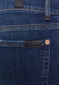 Detailní záběr na kapsu tmavě modrých džínsů se švy v oranžové barvě, černou značkovou nášivkou s nápisem "pro celé lidstvo" a měděným nýtem.